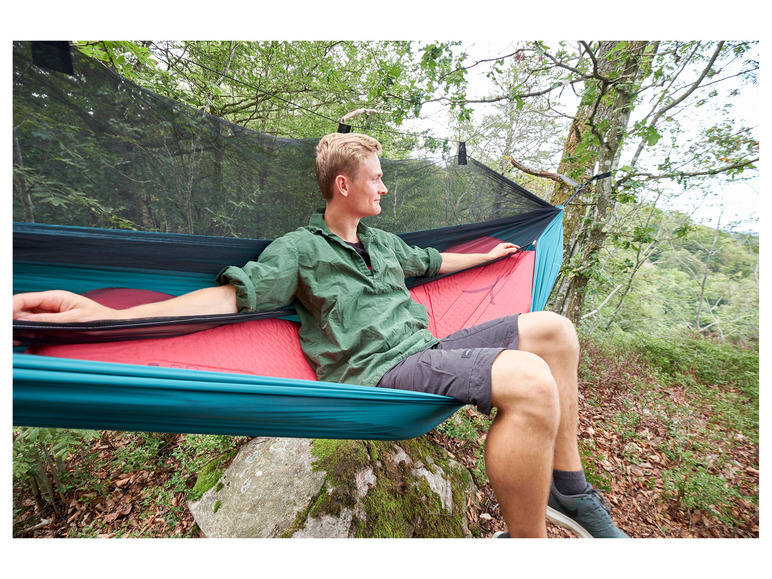 Een man zit in een blauwe hangmat met een rode matras, bevestigd aan twee bomen.