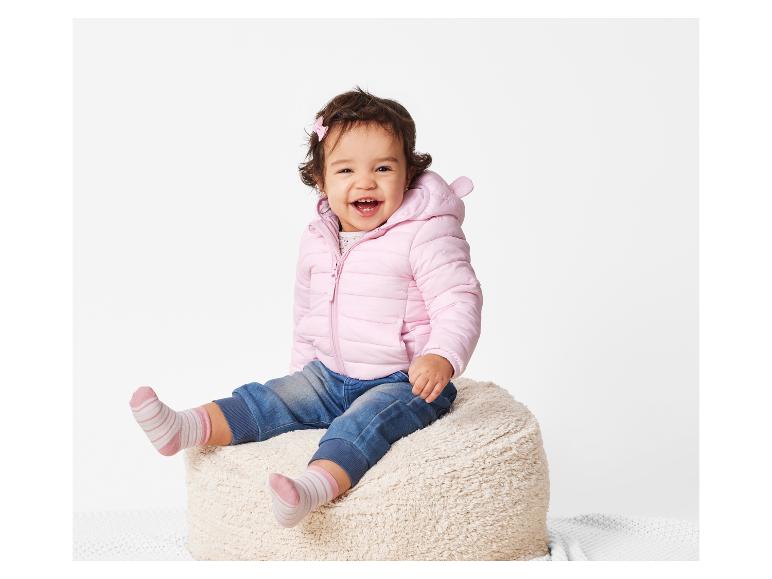 Een lachende baby draagt een roze jasje en gestreepte sokken terwijl hij op een poef zit.