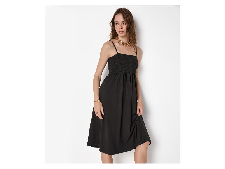 Vrouw in zwarte spaghettibandjesjurk met gouden accessoires, staand bij een witte muur.