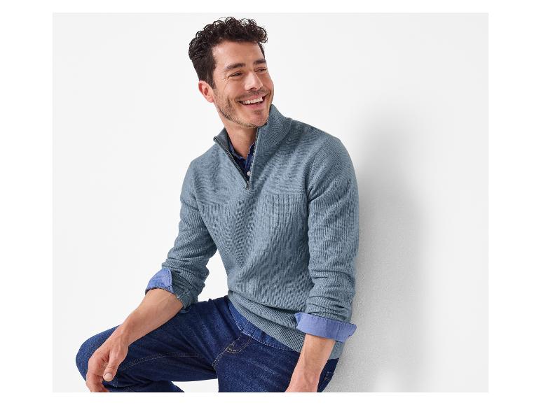 Lachende man in een blauwe trui en jeans, leunend tegen een witte muur.