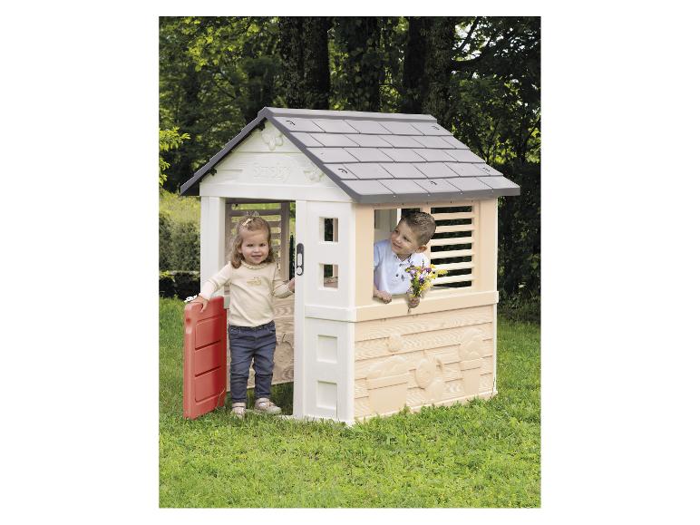 Twee kinderen spelen in een plastic speelhuisje met een grijs dak en rode deur in een tuin.