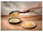 Hand gebruikt crêpemaker om pannenkoeken te maken, met beslag en stapel pannenkoeken op tafel.