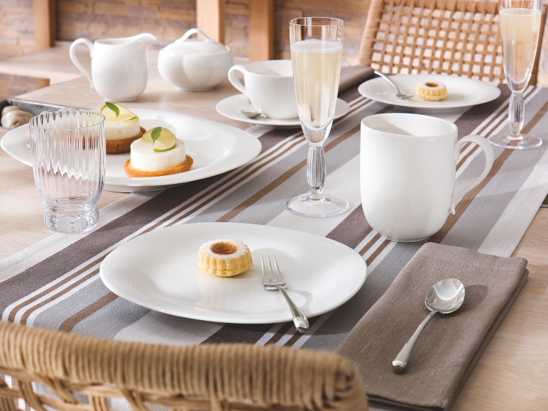 Tafel gedekt met wit porseleinen borden, kopjes, glazen en kleine gebakjes.
