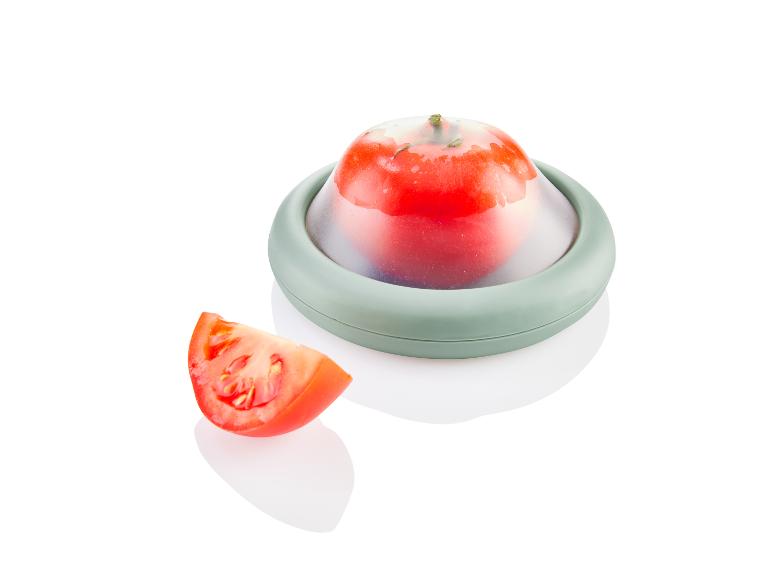 Een groene siliconen deksel over een tomaat, met een gesneden tomaat ernaast.