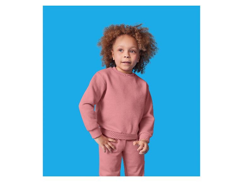 Een jong kind met krullend haar draagt een roze sweatshirt en joggingbroek set.