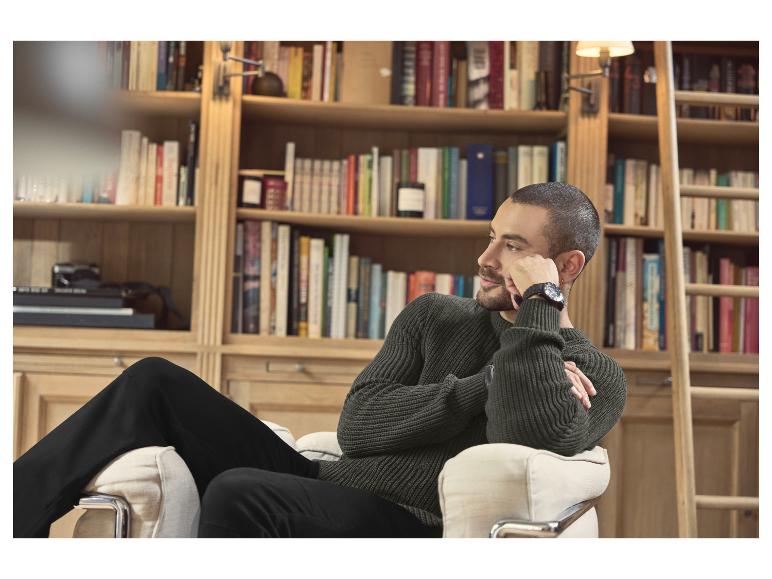 Man in donkergroene gebreide trui en zwarte broek zit in een fauteuil voor een boekenkast.