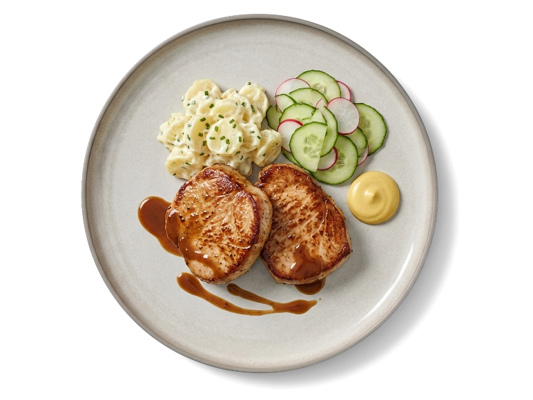 Gebakken varkenshaasmedaillons geserveerd met aardappelsalade, komkommersalade, jus en mosterd.