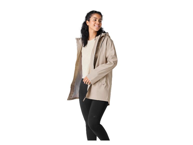 Vrouw in beige jas en zwarte legging, lachend en naar rechts kijkend.
