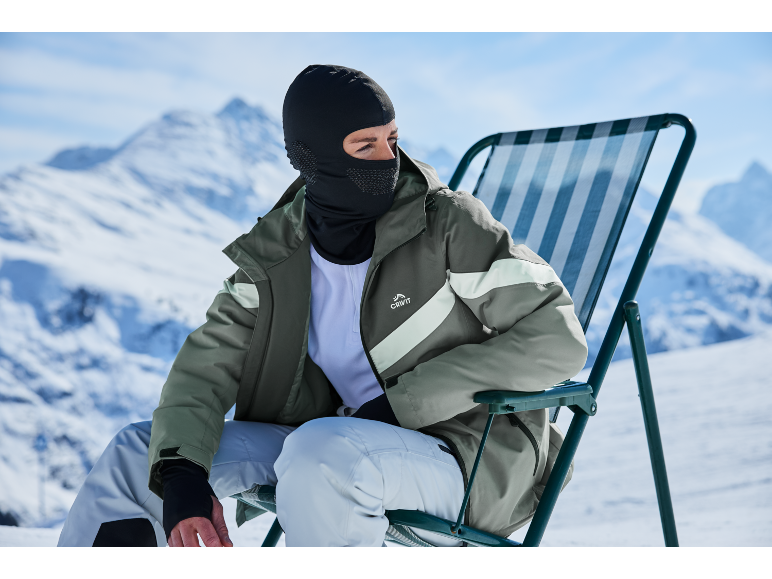 Man met bivakmuts en groene ski-jas zit op een stoel in besneeuwde bergen.