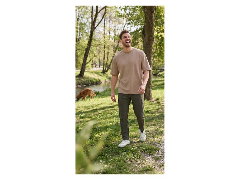 Man in beige t-shirt en groene broek loopt in park, hond op de achtergrond.