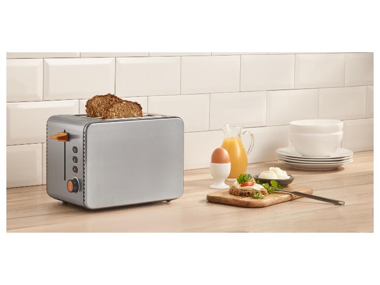 Roestvrijstalen broodrooster met brood, ei, sap en roomkaas op een houten aanrecht.