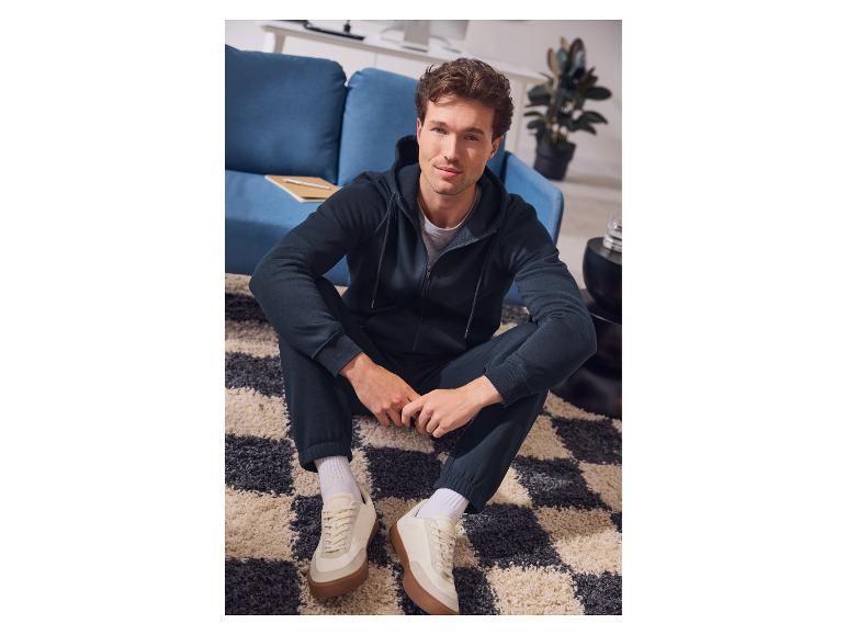 Man in donkerblauwe hoodie en joggingbroek zit op een vloerkleed.