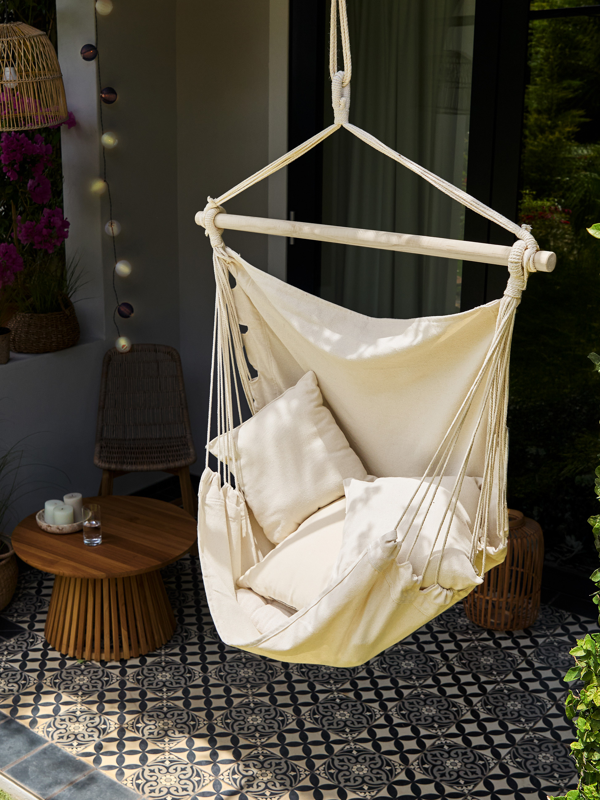 Crèmekleurige stoffen hangstoel met kussens, houten tafel en patroontegels op een balkon.