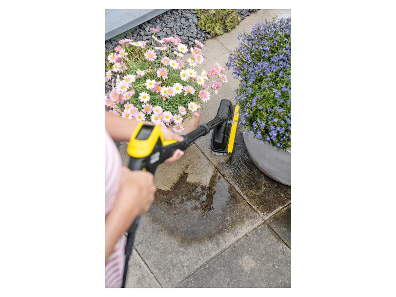Vrouw reinigt terras met hogedrukreiniger en borstel, bloemen op de achtergrond.