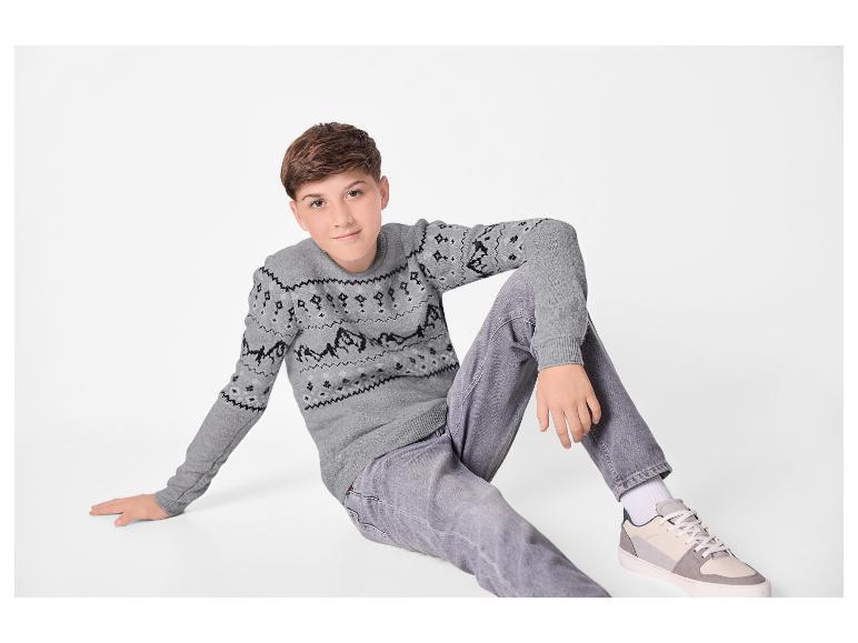Jongen in grijze gebreide trui met patroon en grijze jeans zit op de grond.