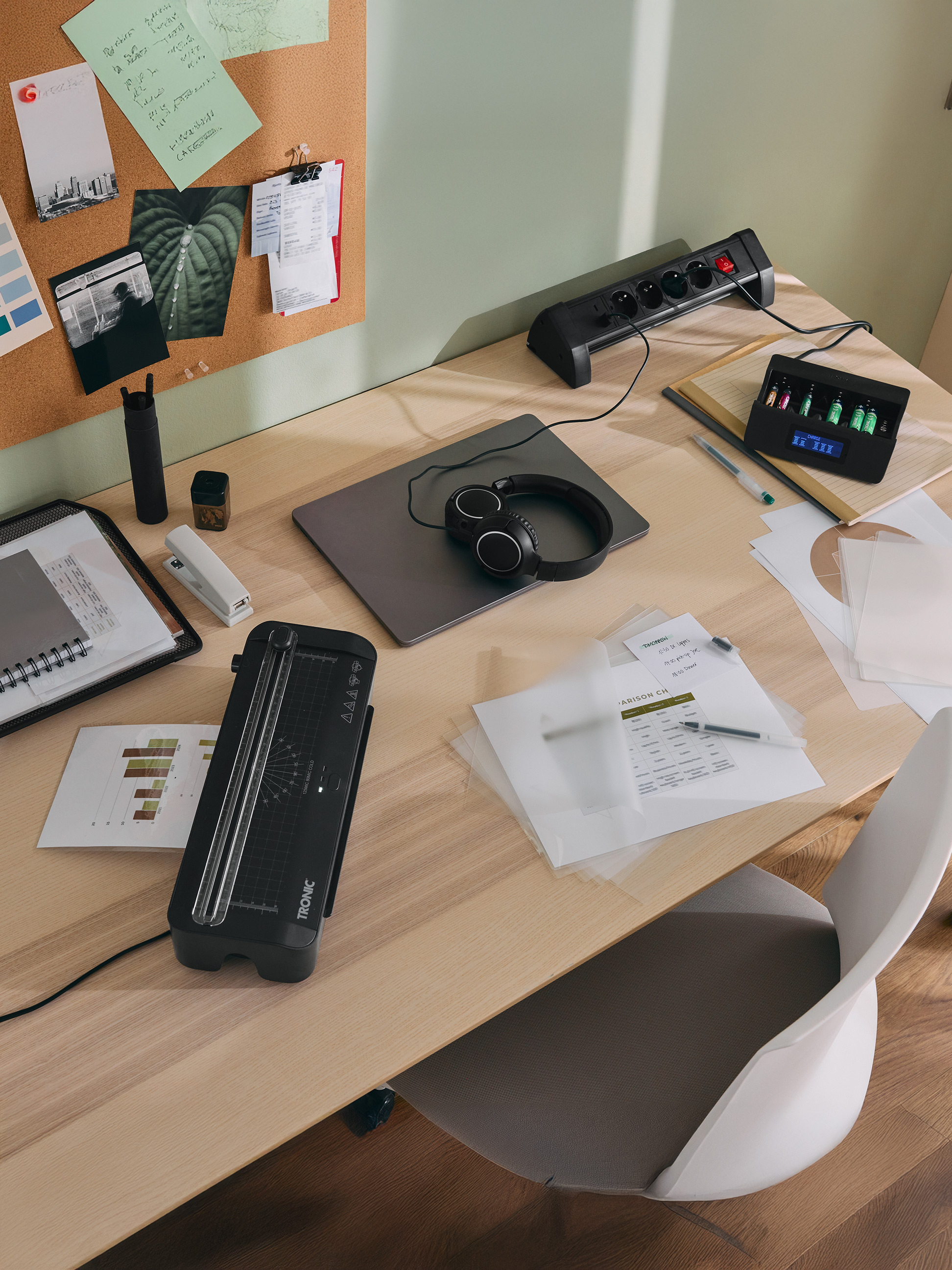 Bureau met TRONIC lamineerapparaat, batterijlader, laptop, koptelefoon en kantoorbenodigdheden.