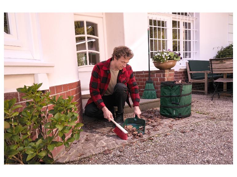 Man veegt bladeren met bezem en blik in groene tuinafvalzak.