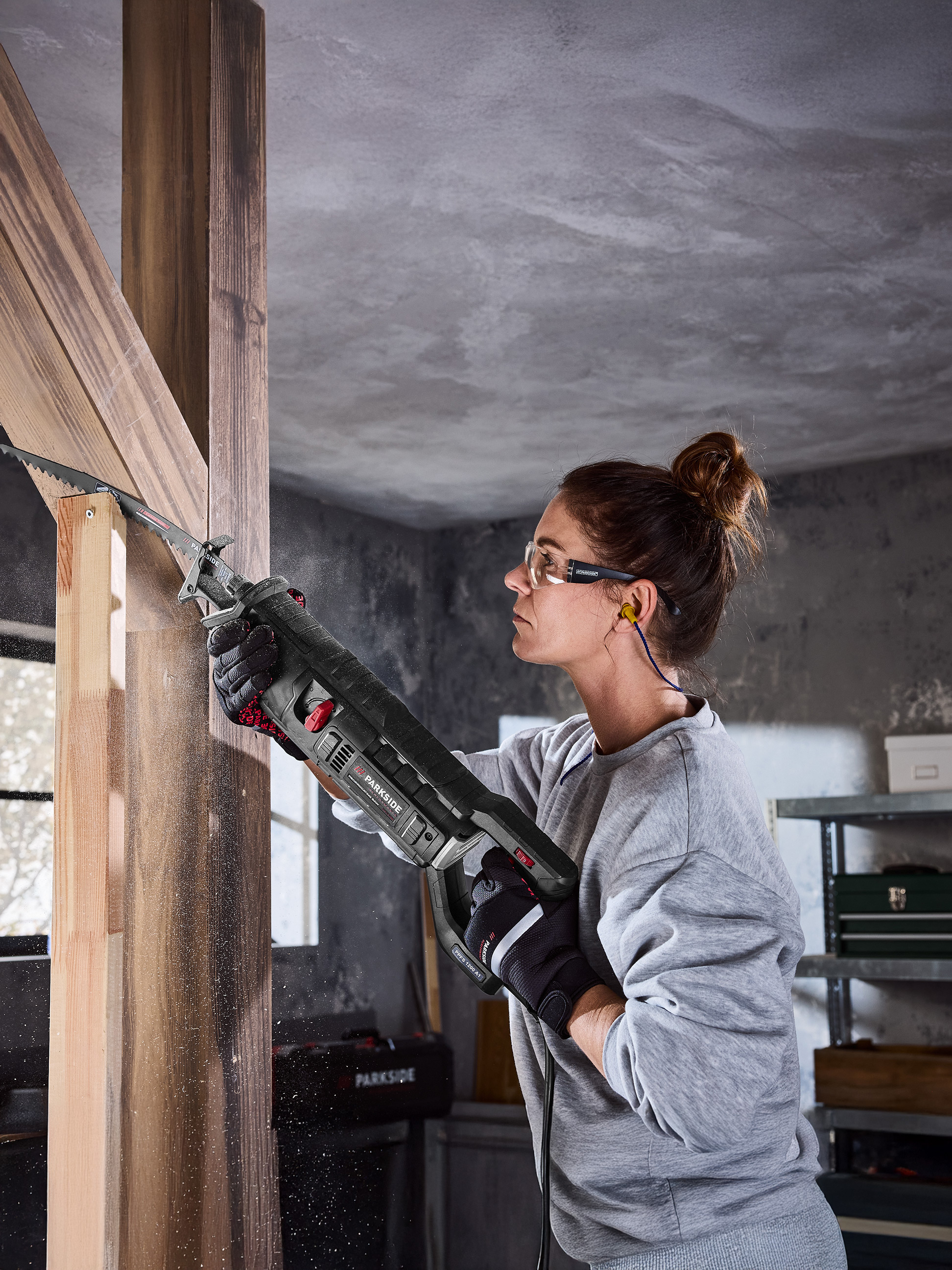 Vrouw gebruikt Parkside reciprozaag om houten balken te zagen, met veiligheidsbril en gehoorbescherming.
