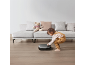 Peuter speelt met een robotstofzuiger op een houten vloer in een woonkamer.