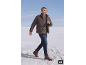 Man in bruine pufferjas, groene trui en jeans, lopend door de sneeuw.