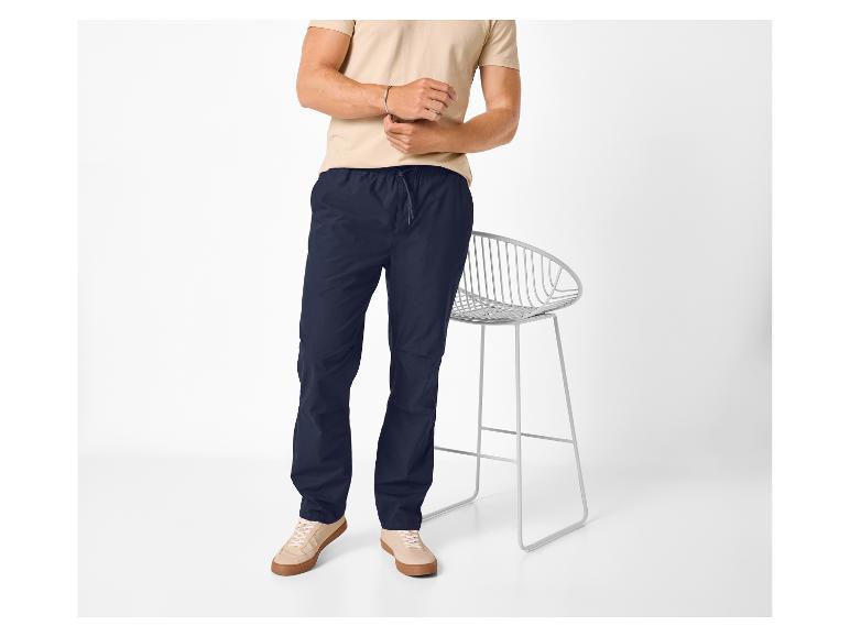 Man in beige T-shirt en donkerblauwe broek, staand naast een stoel.