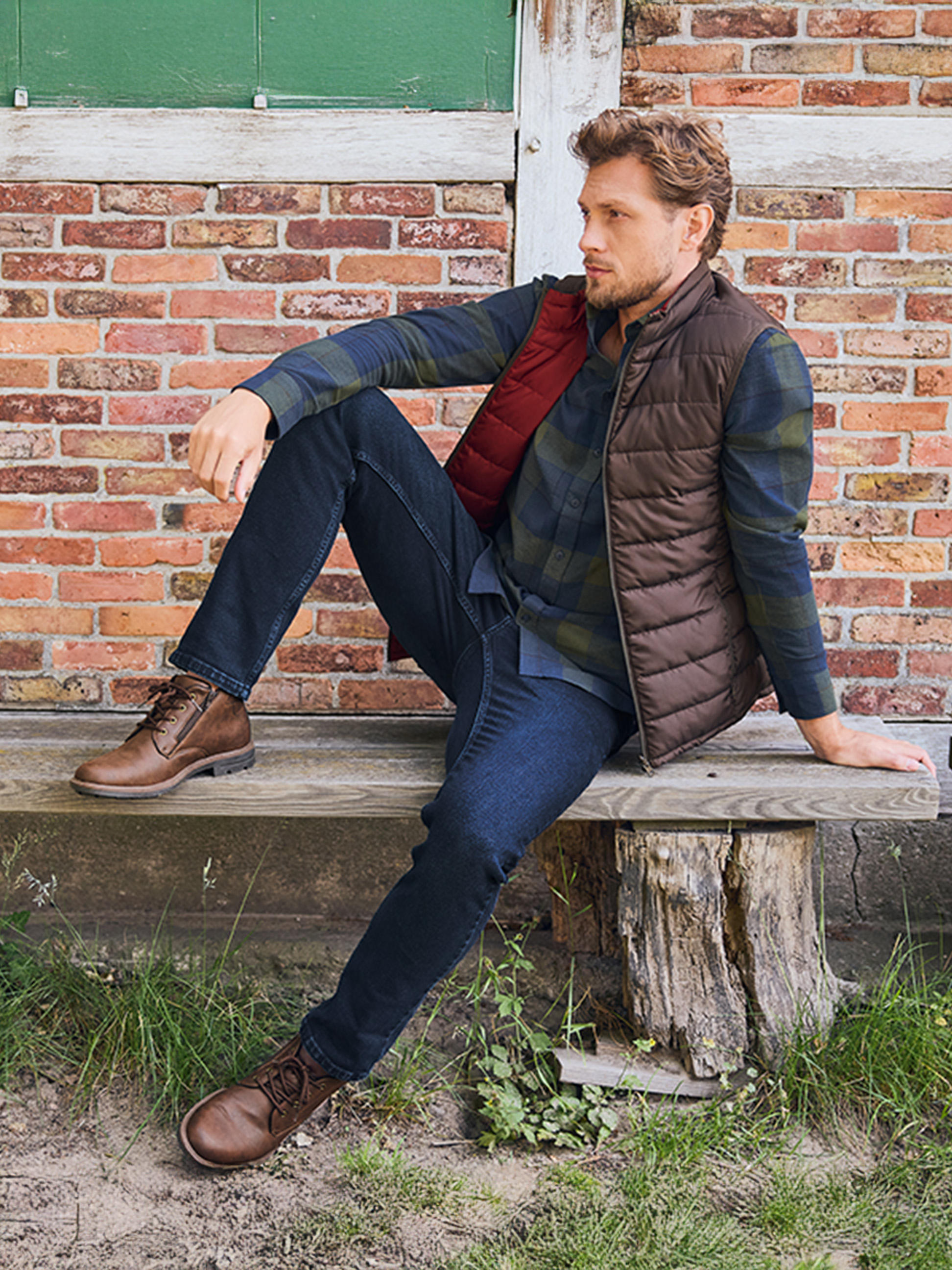 Man in geruit overhemd, gewatteerd vest en jeans zit op een houten bank.