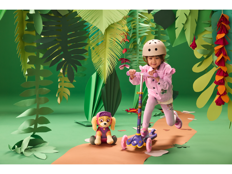 Meisje met helm op een step, naast een knuffelhond, in een jungle-achtige setting.
