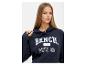 Vrouw in een marineblauwe Bench hoodie met de tekst 'BENCH, 1989, PHYS. ED'.
