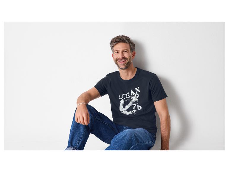 Man in donkerblauw T-shirt met witte anker- en 'OCEAN'-print, blauwe jeans, lachend.