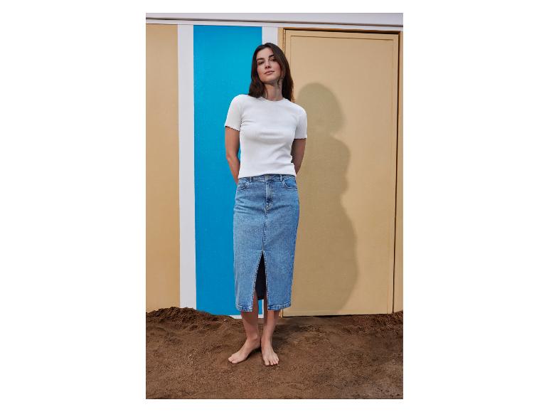 Vrouw in een wit geribbeld T-shirt en een denim midi-rok, blootsvoets staand op zand.