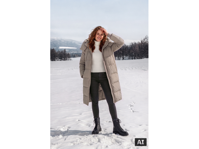 Lachende vrouw in beige lange jas, witte coltrui en zwarte laarzen in de sneeuw.