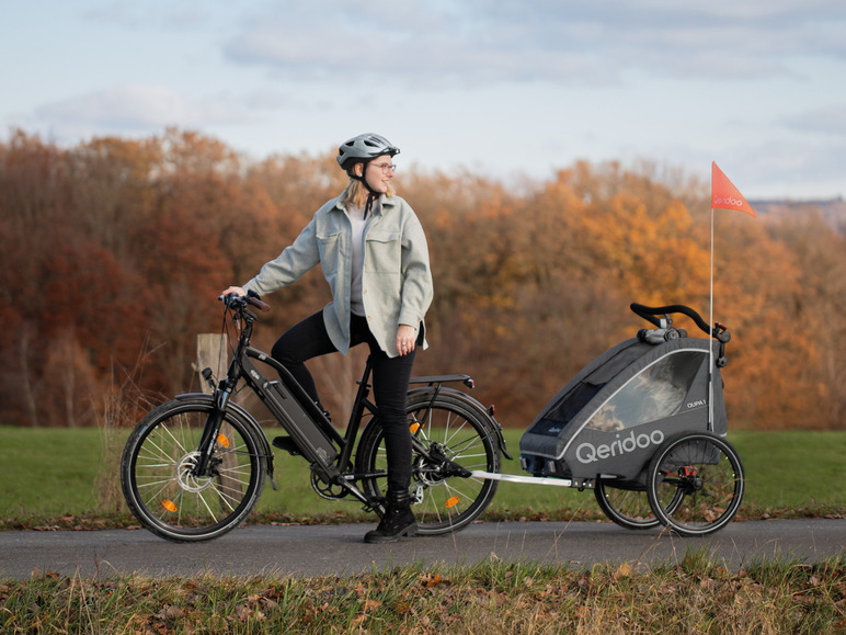 Een vrouw fietst met een elektrische fiets met een Qeridoo kinderwagen erachter.