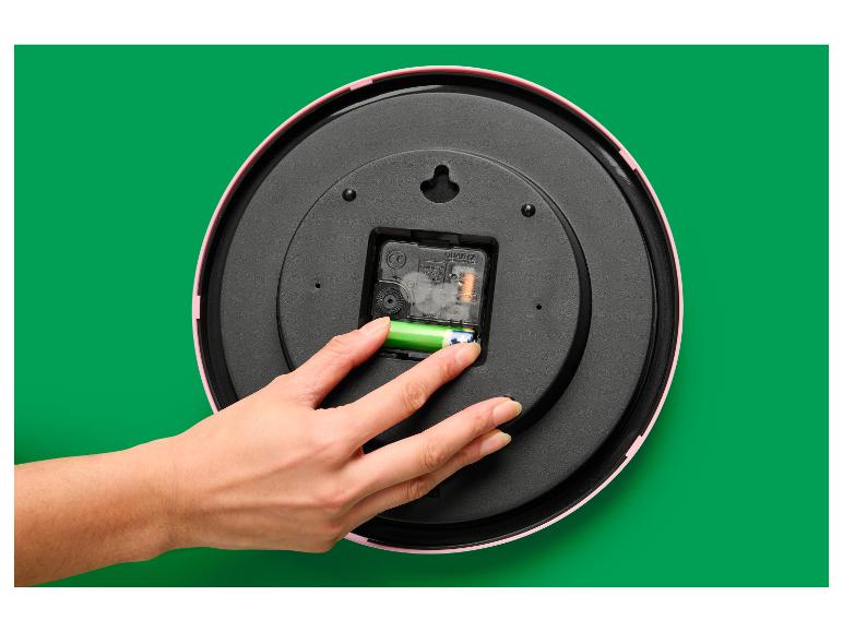 Een hand plaatst een batterij in het mechanisme van een roze wandklok.
