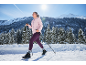 Vrouw in roze jas en paarse legging aan het nordic walken in een besneeuwd landschap.