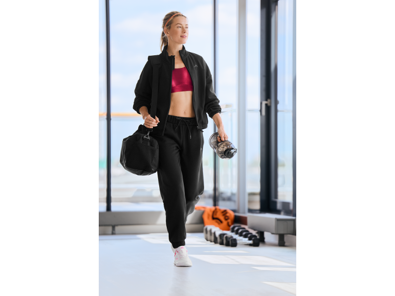 Een vrouw in een zwart trainingspak en rode sportbeha loopt in de sportschool.