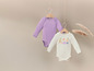 Twee baby-rompers, een in lila en een met een fruitprint, hangen aan houten hangers.
