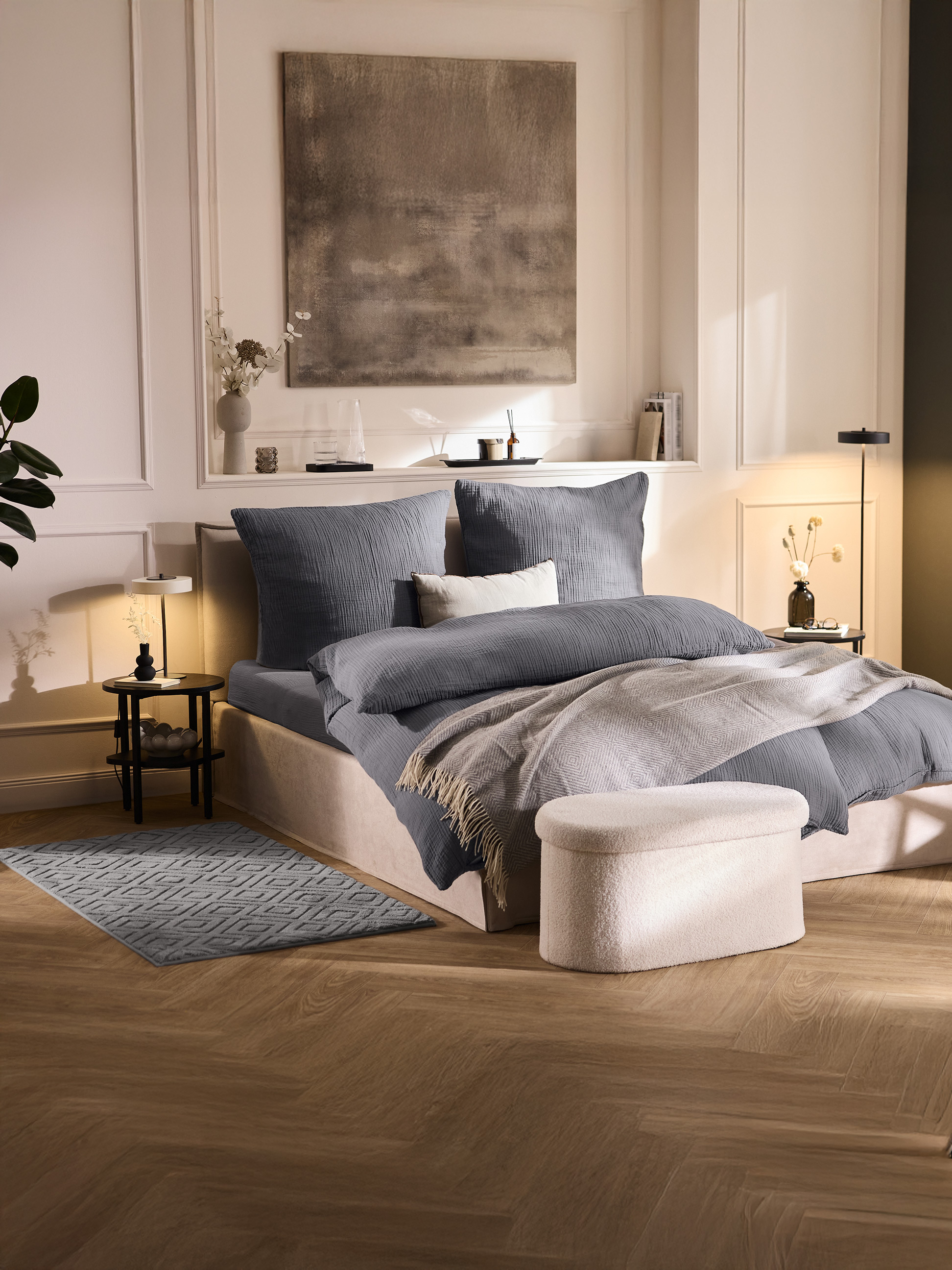 Slaapkamer met een bed met grijs beddengoed, een lichtgrijze plaid en een crèmekleurige poef.
