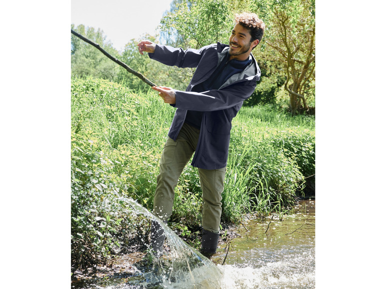 Een man in een blauwe jas en groene broek staat in een beek.