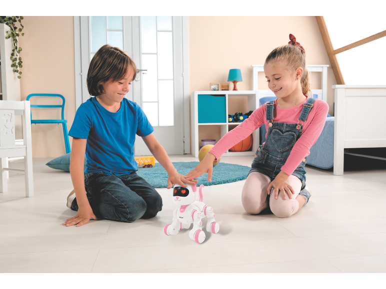 Twee kinderen spelen met een wit en roze robotpuppy op de vloer.