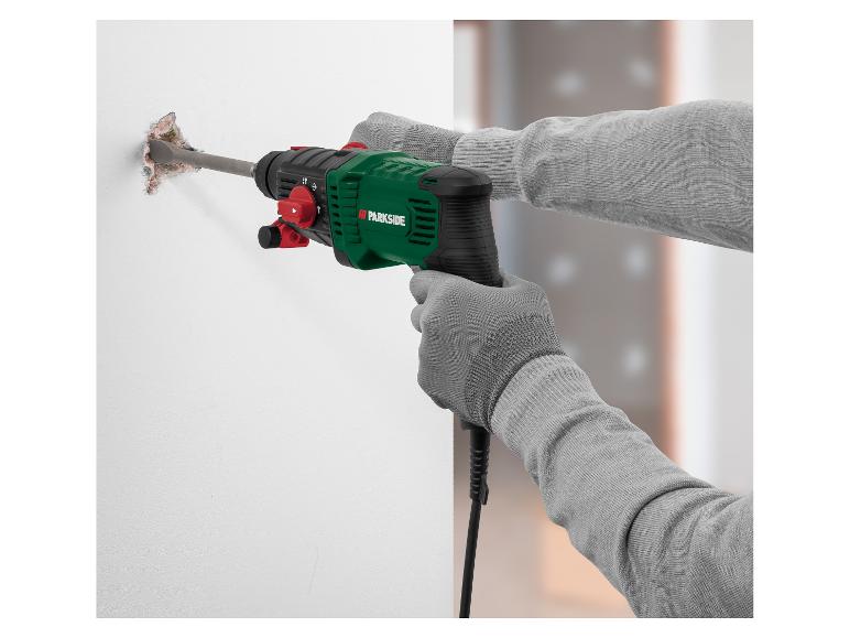 Een man met grijze handschoenen gebruikt een groene Parkside boorhamer om een muur te slopen.
