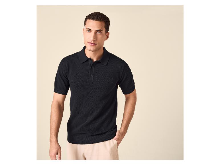 Man in een zwart poloshirt met korte mouwen en een beige broek, poserend met hand in zak.