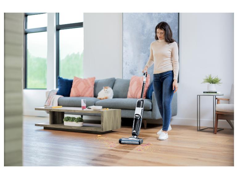 Vrouw maakt houten vloer schoon met een Bissell CrossWave vloerreiniger, terwijl een kat op de bank ligt.