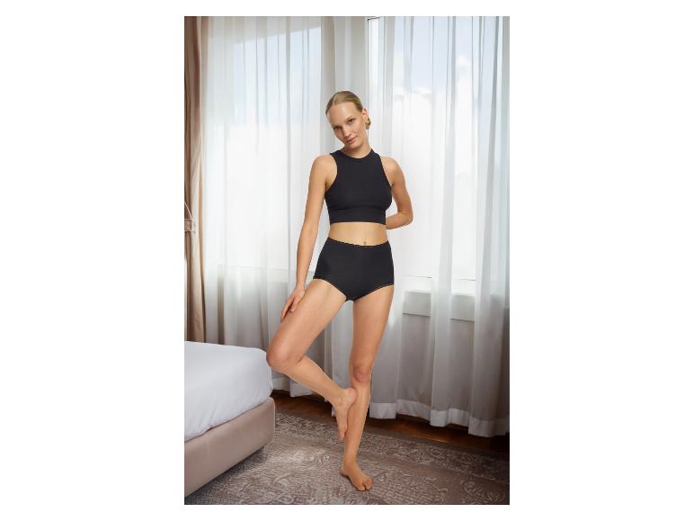Vrouw in zwart ondergoed, crop top en hoge taille slip, poserend in een slaapkamer.