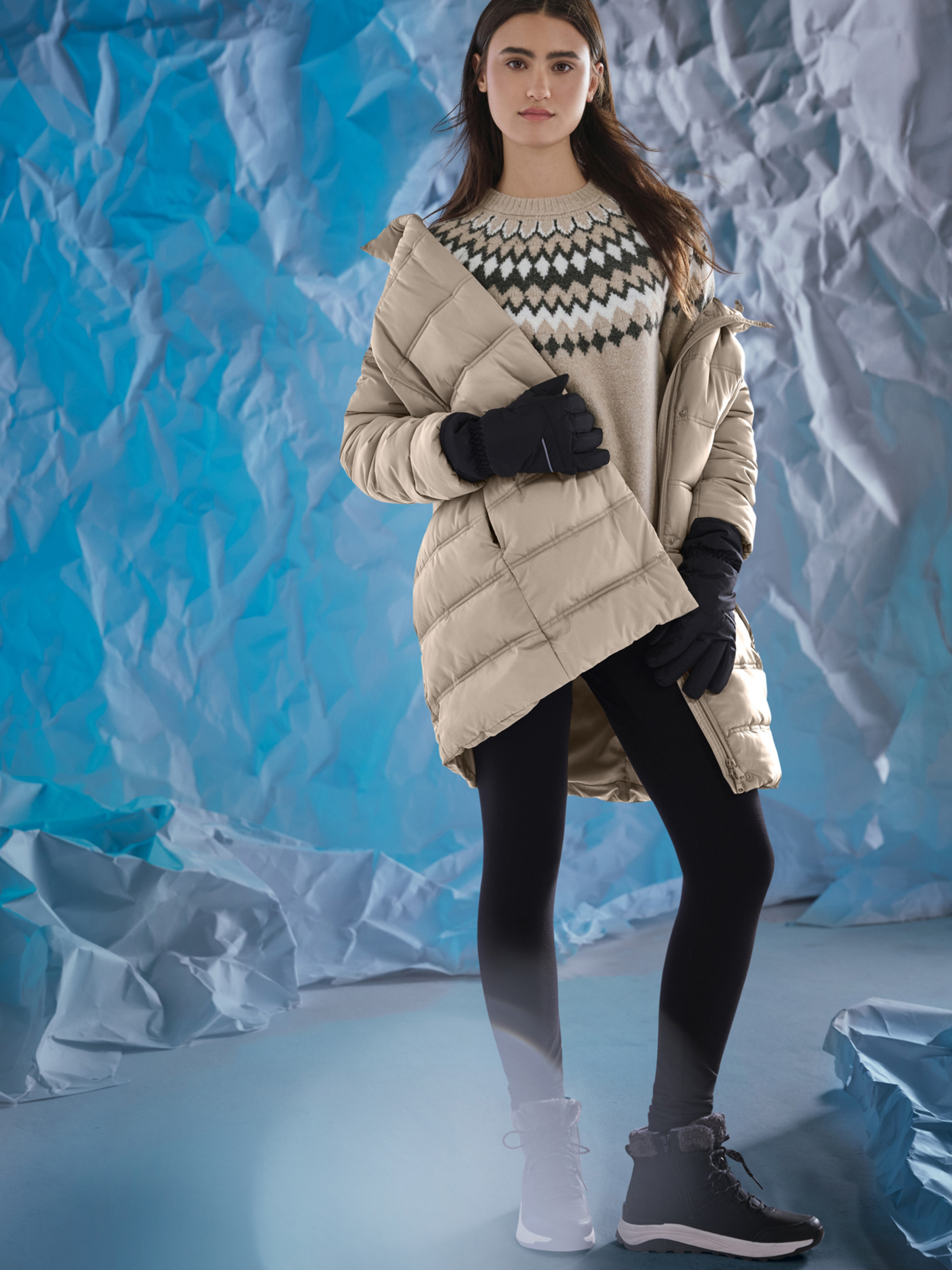 Vrouw in beige jas, patroon trui, zwarte legging en winterlaarzen, tegen een blauwe achtergrond.