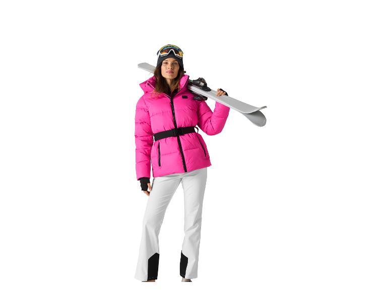 Vrouw in roze ski-jas, witte broek en skibril, met ski's.