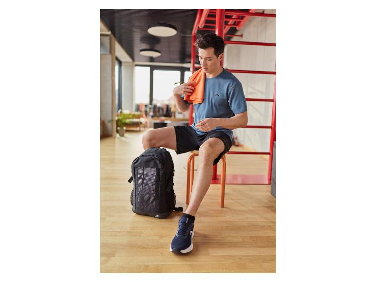 Man in sportshirt en korte broek zit naast rugzak, met oranje handdoek.