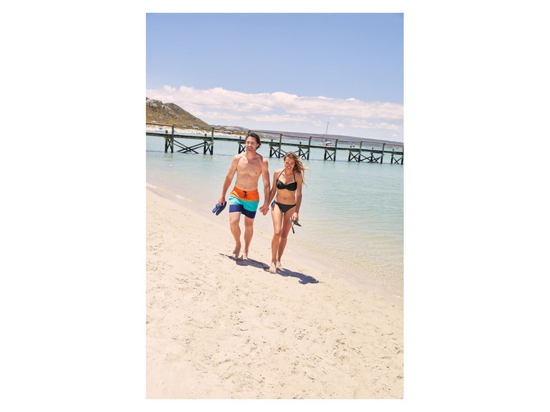 Een stelletje loopt hand in hand over een strand met zwemkleding aan.