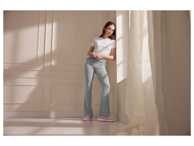 Vrouw in een wit T-shirt en grijze flared leggings, leunend tegen een muur.
