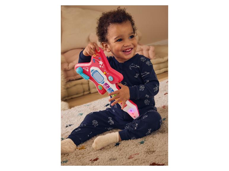 Een lachende baby in een blauwe onesie speelt met een roze speelgoedgitaar.