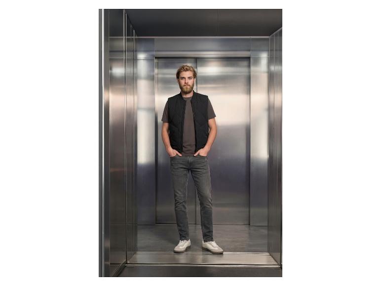 Man in een vest in een lift, draagt jeans en een t-shirt.